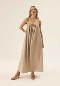 Beżowa długa sukienka z regulowanymi ramiączkami, marszczonym dekoltem i zwiewną sylwetką. Połączona z brązowymi sandałami na obcasie.
