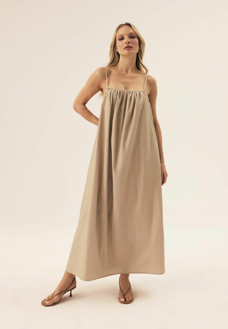 Beżowa długa sukienka z regulowanymi ramiączkami, marszczonym dekoltem i zwiewną sylwetką. Połączona z brązowymi sandałami na obcasie.