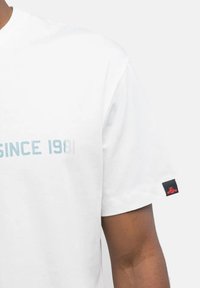 Camiseta de algodón blanca con gráfico en texto azul claro "DESDE 1981" y una etiqueta negra con emblema rojo en la manga izquierda.