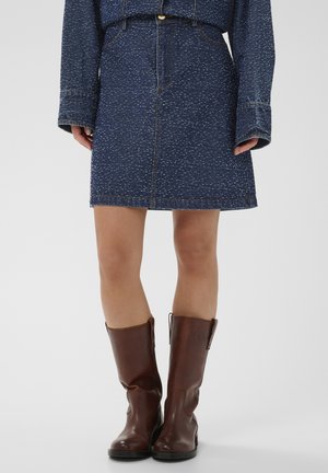 Denimnederdel med et mørkeblåt plettet mønster, knapper foran og lynlås. Kombination med brun læderstøvler til knæene.