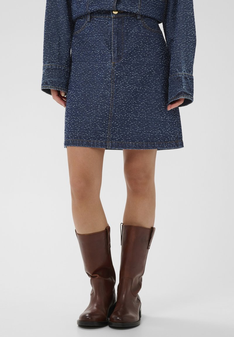 Fustă din denim cu un model pătat albastru închis, cu nasturi în față și închidere cu fermoar. Asortată cu cizme maro din piele până la genunchi.