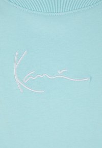 Light blue fabric featuring a white embroidered signature design. Smooth texture with a ribbed neckline. Minimalistic and modern appearance.