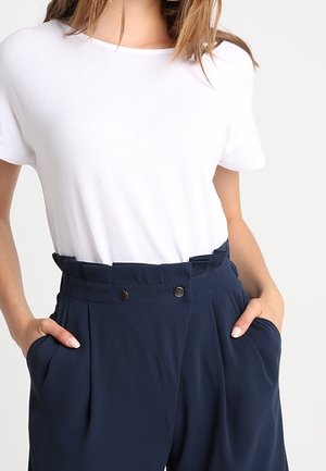 Nainen pukeutuneena yksinkertaiseen valkoiseen lyhythihaiseen paitaan, joka on laitettu korkeavyötäröisten, laivastonsinisten pliseerattujen housujen sisään, kädet taskuissa.