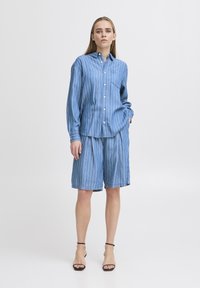 Camicia a righe blu con bottoni e maniche lunghe, dotata di tasca, abbinata a pantaloni corti plissettati blu. Indossata con sandali neri con cinturini.