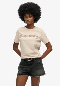 Beige bomuld t-shirt med "Superdry" logo i brunt, parret med sorte denim shorts med frynset kant og et sort bælte med en sølvspænde.