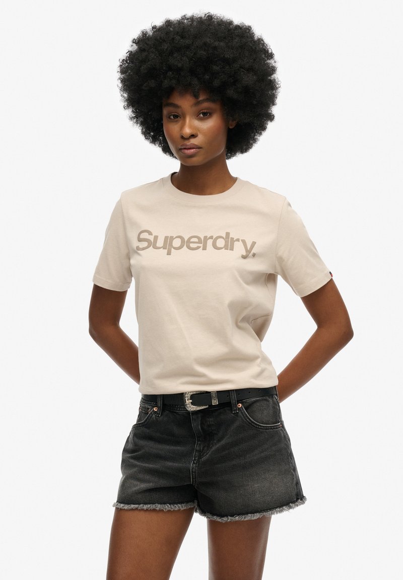 Beige bomuld t-shirt med "Superdry" logo i brunt, parret med sorte denim shorts med frynset kant og et sort bælte med en sølvspænde.