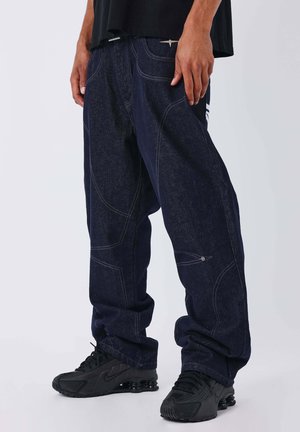 Donkerblauwe denim broek met een losse pasvorm, contrasterende stiksels en zijzakken. Gekleed met zwarte sportschoenen, die een casual ontwerp tonen.