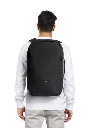 Uomo che indossa uno zaino Bellroy nero con spallacci imbottiti, vestito con una felpa bianca e pantaloni grigi chiari, girato di spalle su uno sfondo bianco.