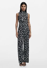 Svart jumpsuit med hög krage och vida ben, prydd med ett vitt prickigt mönster. Tillverkad av ett mjukt, lätt tyg.