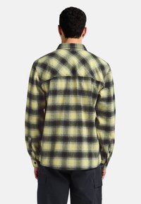 Chemise en flanelle avec un motif à carreaux jaunes et noirs, manches longues, poignets à boutons, et un ourlet arrondi avec une pièce de dos.