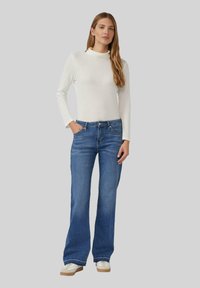 Un haut blanc à manches longues, ajusté, associé à un jean bleu évasé. Les jeans possèdent des poches et une fermeture à bouton. Texture de tissu lisse.