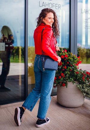 Red leather cropped jacket, blue denim jeans, black sneakers, and gray crossbody bag with "VUCH" embossed. Flower pot in background.