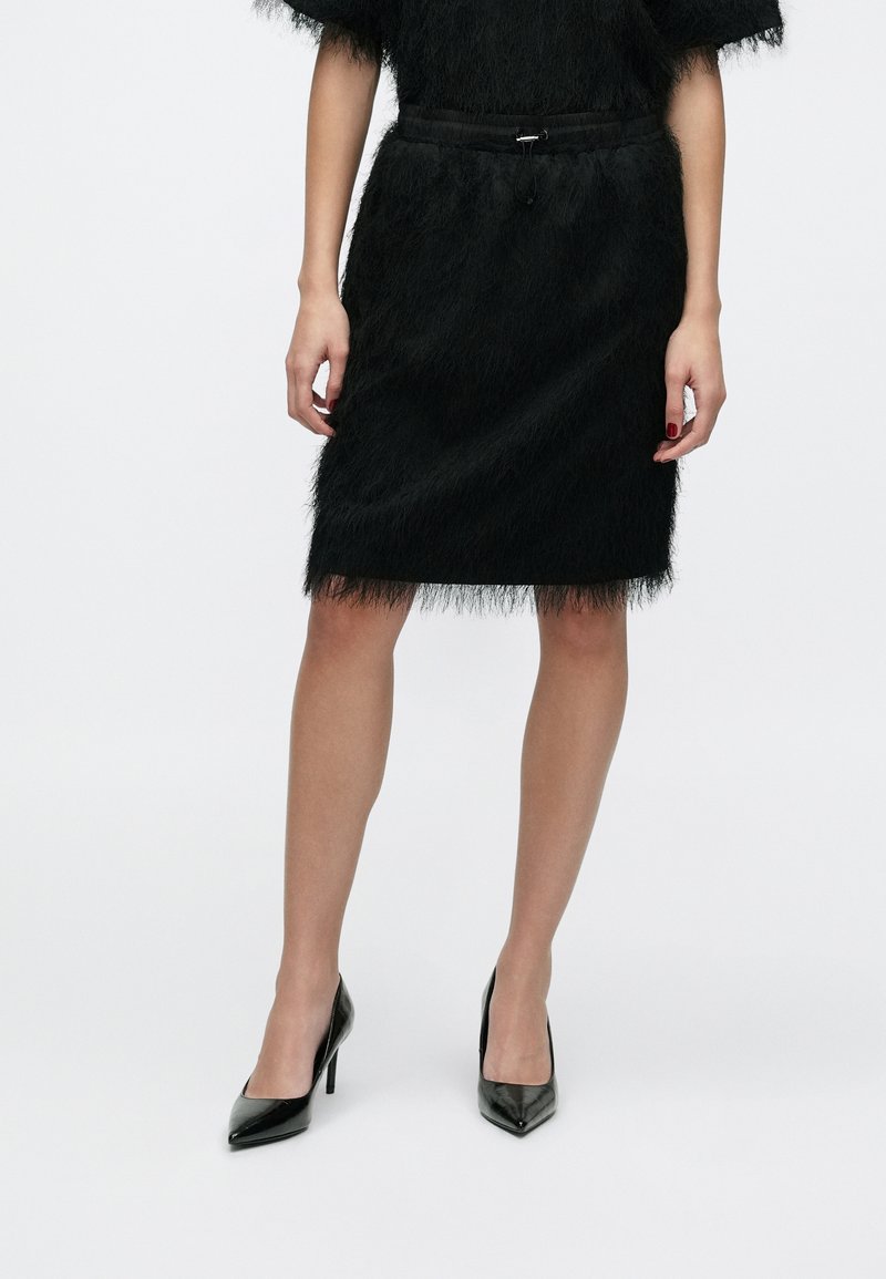 Czarna spódnica wykonana z teksturowanego, puszystego materiału, uzupełniona dopasowaną sylwetką, taliowaniem i zestawiona z błyszczącymi czarnymi szpilkami.