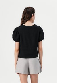 Top nero con maniche a palloncino, texture uniforme, scollo rotondo e design aderente. Abbinato a pantaloni corti grigi chiari, entrambi mostrati di spalle.