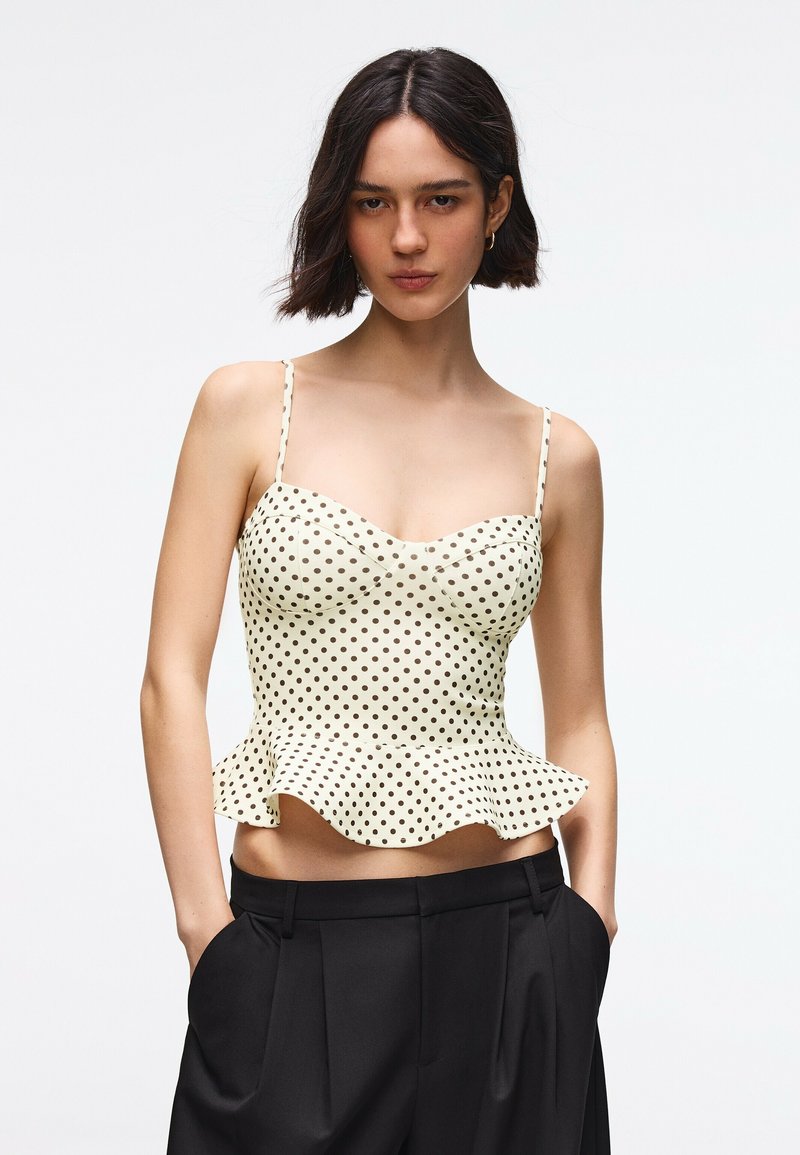 Femme portant un top sans manches blanc à pois noirs avec un ourlet volanté et un pantalon noir taille haute, mains dans les poches, fond neutre.