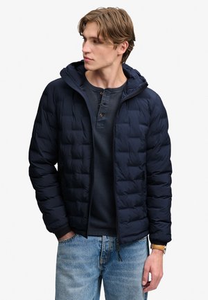 Young man wearing a navy quilted jacket with hood, dark henley shirt, light blue jeans, and a brown leather watch, looking to his left.