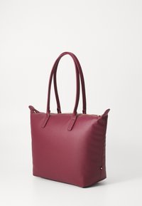 Sac fourre-tout bordeaux en matériau synthétique lisse, doté de deux poignées allongées, d'une fermeture éclair et d'un petit logo sur le côté.