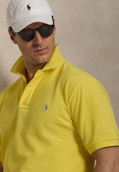 Homme portant des lunettes de soleil noires, une casquette de baseball blanche avec un logo, et un polo jaune avec un petit logo brodé sur la poitrine gauche.