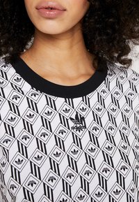 Nahaufnahme einer Frau in einem schwarz-weißen Adidas-Shirt mit einem sich wiederholenden Trefoil-Logo-Muster und schwarzem Kragen.