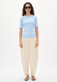 Helles blaues, geripptes T-Shirt mit mittellangen Ärmeln, kombiniert mit cremefarbenen, weiten Hosen. Schwarze Flip-Flops und dunkle Sonnenbrillen runden den Look ab.