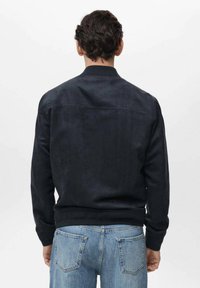 Chaqueta bomber de ante negro con cuello y puños acanalados, que presenta una textura simple y suave, llevada sobre unos jeans de azul claro.