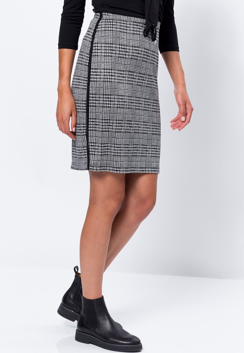 Jupe à carreaux noir et blanc avec un design ajusté, une coupe jusqu'au genou et des détails sur les coutures latérales. Associée à des bottines noires.