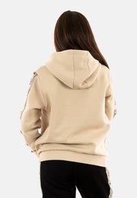 Felpa beige con cappuccio, texture liscia, polsini e orlo a costine, con dettagli decorativi lungo le maniche, abbinata a pantaloni neri.