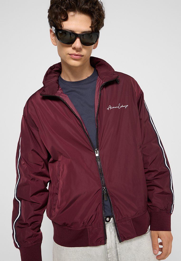 Giacca bomber bordeaux con chiusura a zip, polsini e orlo a coste. Presenta strisce laterali bianche e ricamo del logo sul petto. Tessuto dalla texture liscia.