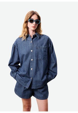 Femme portant une chemise bleu à fines rayures boutonnée et un short assorti, avec de grandes lunettes de soleil bleues, les mains dans les poches.
