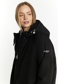 Giacca nera impermeabile con cappuccio rimovibile, zip frontale e polsini elasticizzati. Logo DRI-MASTER ben visibile sulla manica destra.