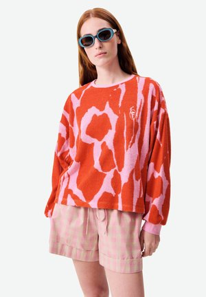 Femme aux longs cheveux roux portant des lunettes de soleil bleues, un pull surdimensionné à motif orange et rose, et un short à carreaux rose clair.