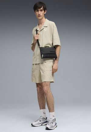 Sac bandoulière noir à texture lisse avec une sangle ajustable, accompagné d'une chemise beige à manches courtes et d'un short cargo beige.