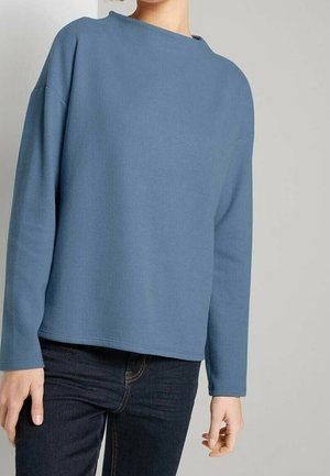 Person trägt ein blaues, langärmeliges Rundhalsoberteil und dunkle Jeans, steht vor einem einfarbigen hellgrauen Hintergrund.