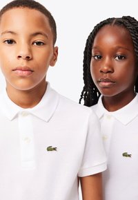 Twee kinderen in witte poloshirts met groene krokodillenlogo's, dichtbij tegen een effen witte achtergrond staand.