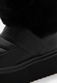 Černé platformové boty s lesklým koženým základem a černým umělým kožešinovým svrškem. Na popruhu je vyražené logo "Love Moschino".