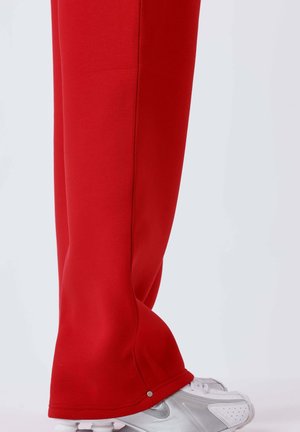 Pantalon rouge à jambes larges couvrant des baskets blanches avec le logo Nike argenté, vu de côté sur un fond clair uni.