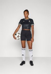 Juoda trumpa sportinė marškinė su lapų raštais, derinama su juodomis šortais, baltomis kojinėmis iki kelių su juodomis juostelėmis ir juodais futbolo batais, laikant kamuolį.
