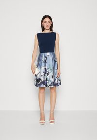A sleeveless dress with a fitted navy top and a flowy skirt featuring a floral pattern in shades of blue and white, paired with strappy white heels.
