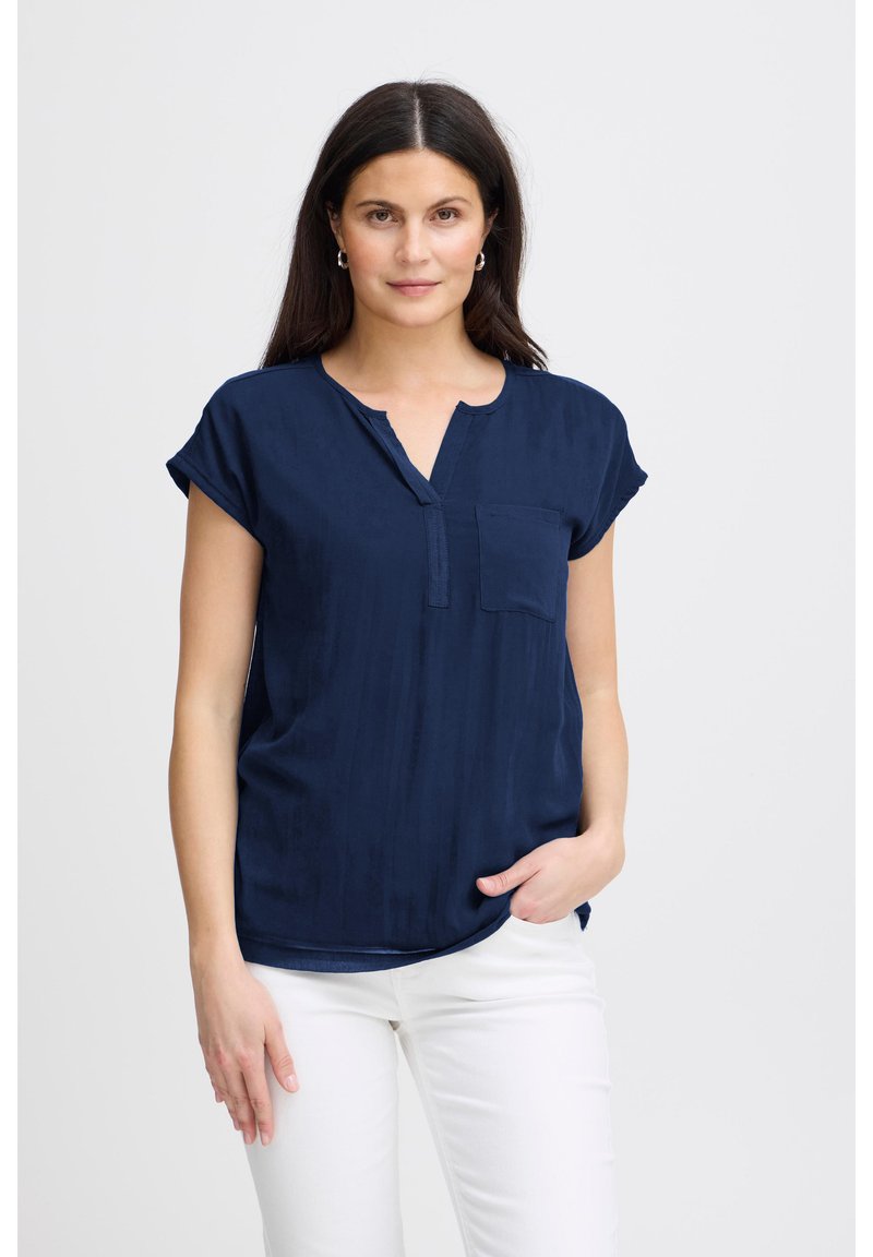 Femme aux cheveux foncés portant un chemisier bleu marine à manches courtes et un pantalon blanc, debout avec une main dans la poche devant un fond uni.
