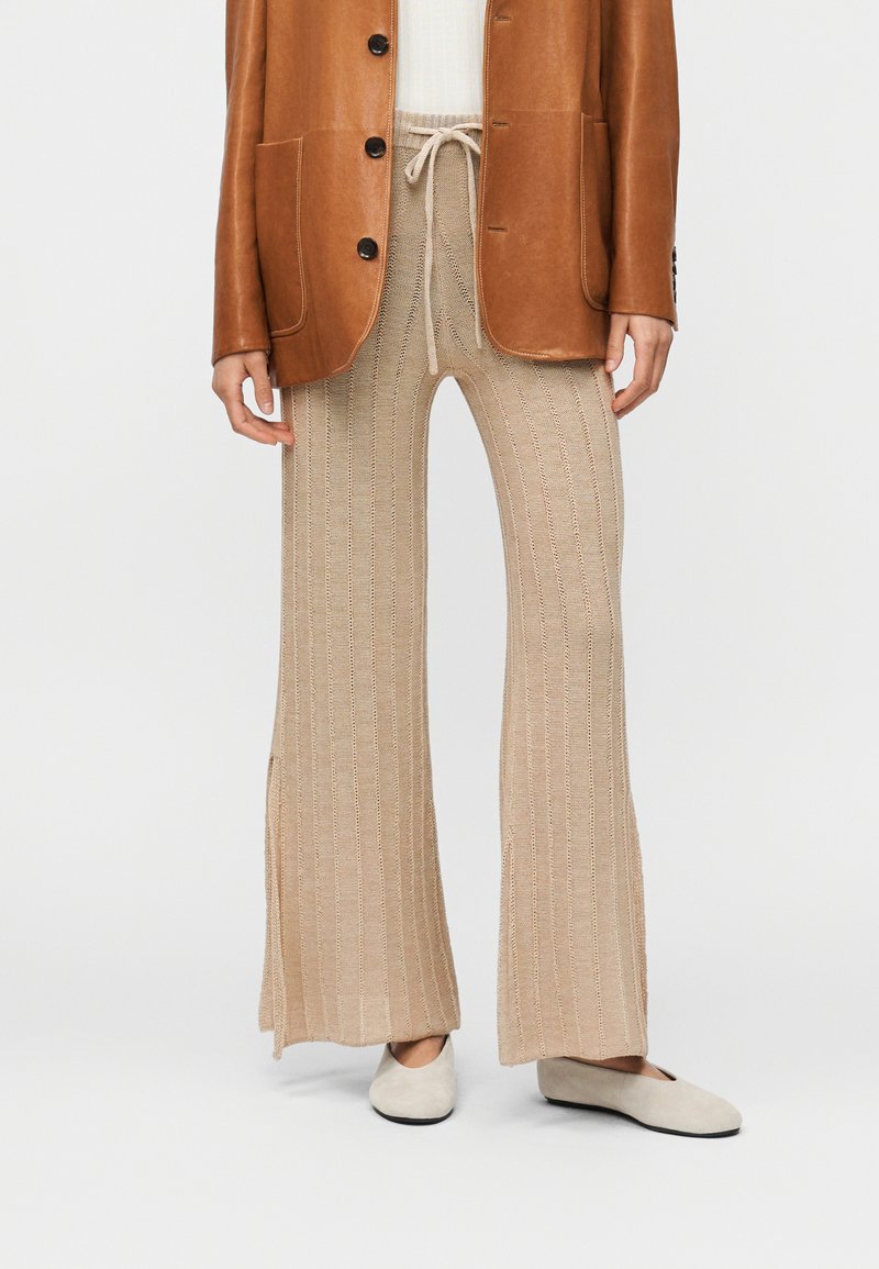 Μπεζ πλεκτό παντελόνι με φαρδιά πόδια και πλαϊνές σχισμές, με κορδόνι στη μέση, συνδυασμένο με καφέ δερμάτινο μπουφάν και κρεμ επίπεδες παπούτσια.