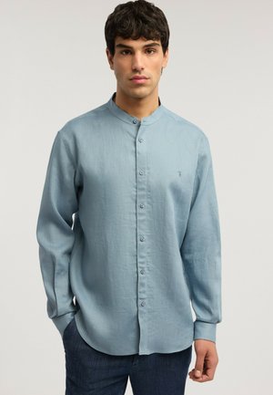 Mann mit dunklem lockigem Haar, der ein hellblaues Hemd mit Stehkragen und Knöpfen sowie dunkle Jeans trägt, vor einem einfarbigen Hintergrund steht.