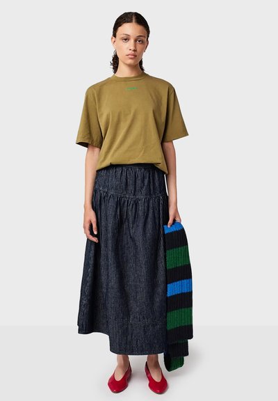 Una donna con i capelli ricci indossa una maglietta verde oliva, una lunga gonna scura a righe, tiene una sciarpa a righe blu e verde e indossa delle ballerine rosse.