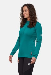 Türkiser Pullover mit langen Ärmeln, halbem Reißverschluss und strukturierter Oberfläche. Merkmale sind ein Logo am Ärmel und ein Etikett am Saum. Getragen mit schwarzen Hosen.