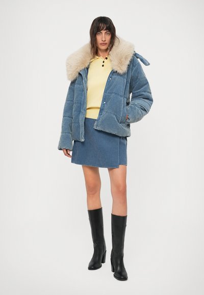 Kvinde iført en blå kordfløjl pufferjakke med pelskant, matchende nederdel, gul sweater og sorte højhælede støvler til knæene, poserer stående.