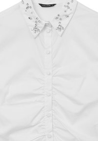 Camicia bianca con design aderente, colletto decorato con strass trasparenti, chiusura frontale con bottoni e texture arricciata lungo i lati.