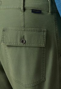 Zelené kalhoty s zadní kapsou na knoflíkovém zapínání a švy. Materiál má hladkou texturu s jemným leskem.