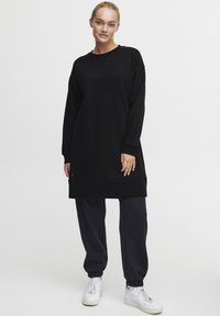 Svart oversized sweatshirtklänning med långa ärmar, ribbade ärmslut och en nederdel som går till mitten av låret, i kombination med marinblå joggers och vita sneakers.