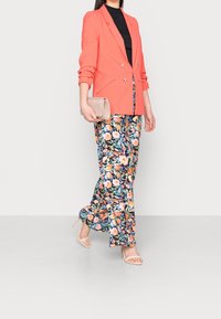 Blazer doppio petto corallo, top nero, pantaloni a gamba larga floreali e tacchi beige con cinturini, abbinati a una borsa a mano rosa lucida.