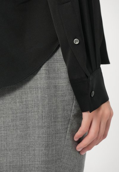 Nahaufnahme einer Hand, die seitlich ruht, wearing ein schwarzes Langarmhemd mit Knöpfen und graue strukturierte Hose.