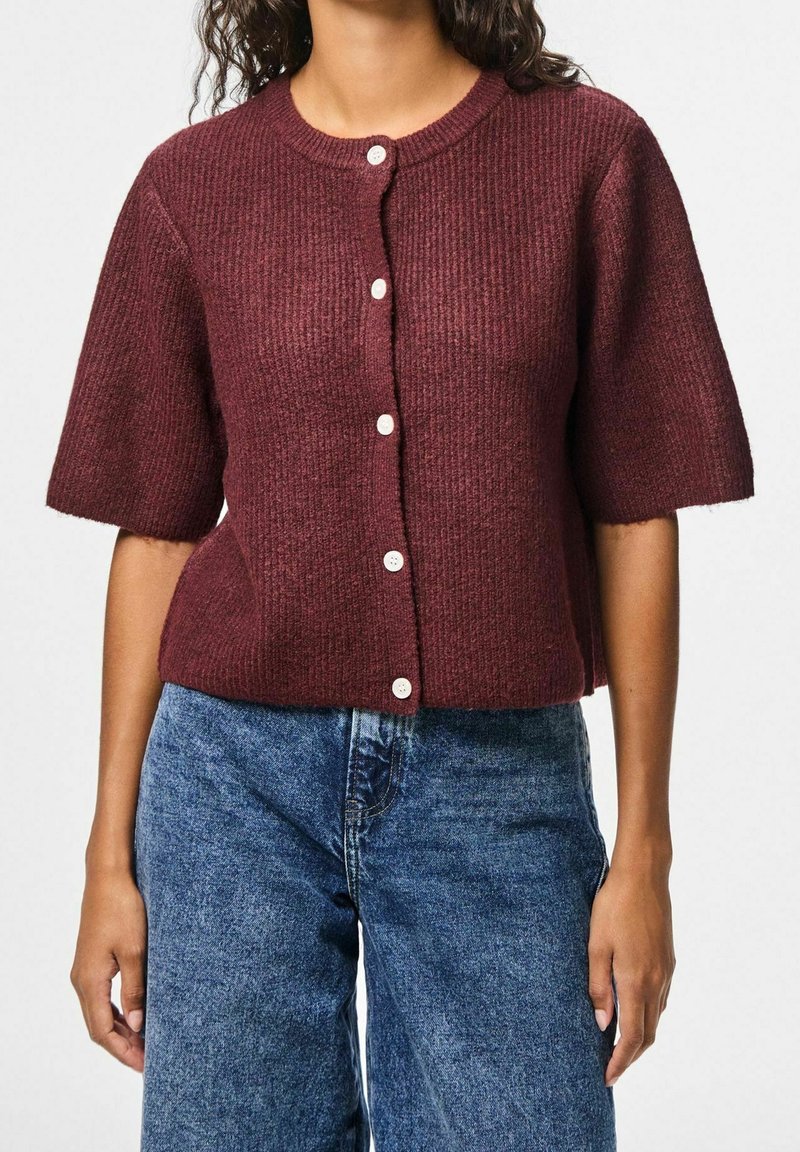 Femme portant un cardigan côtelé bordeaux à manches courtes avec des boutons blancs, associé à un jean bleu taille haute.